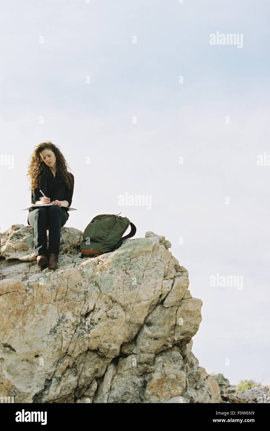 Woman Rocks Writing A woman sitting on a rock, writing in a notebook, a backpack lying by ...