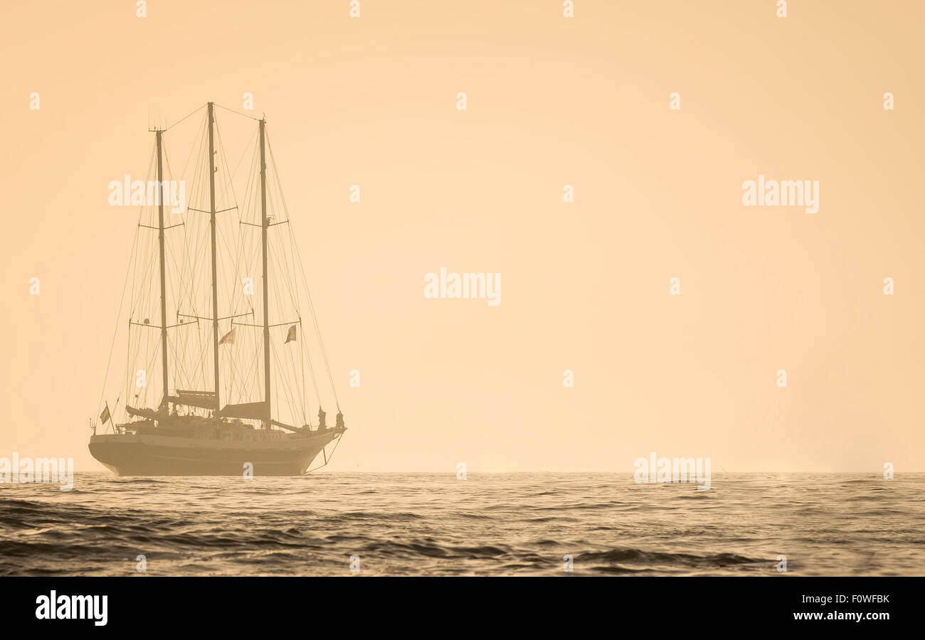 A sailboat travels at sea to the horizon. It is fogy and the sun goes down. Stock Photo