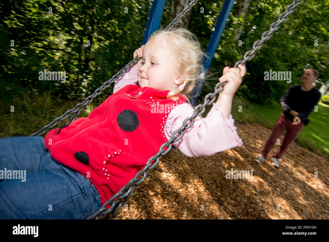 Little girl on swing with Dad in background. Stock Photo