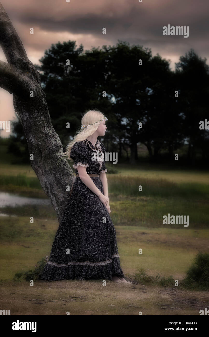 a woman in a black dress with flower in her hair is leaning against a tree Stock Photo