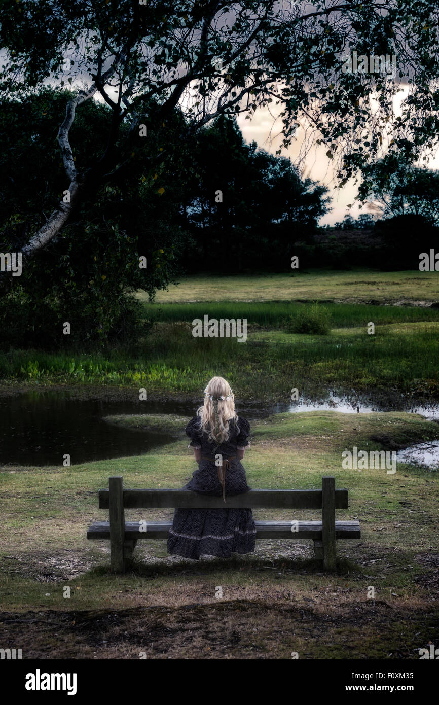 a blonde girl in a floral dressl is sitting on a bench at a pond Stock Photo