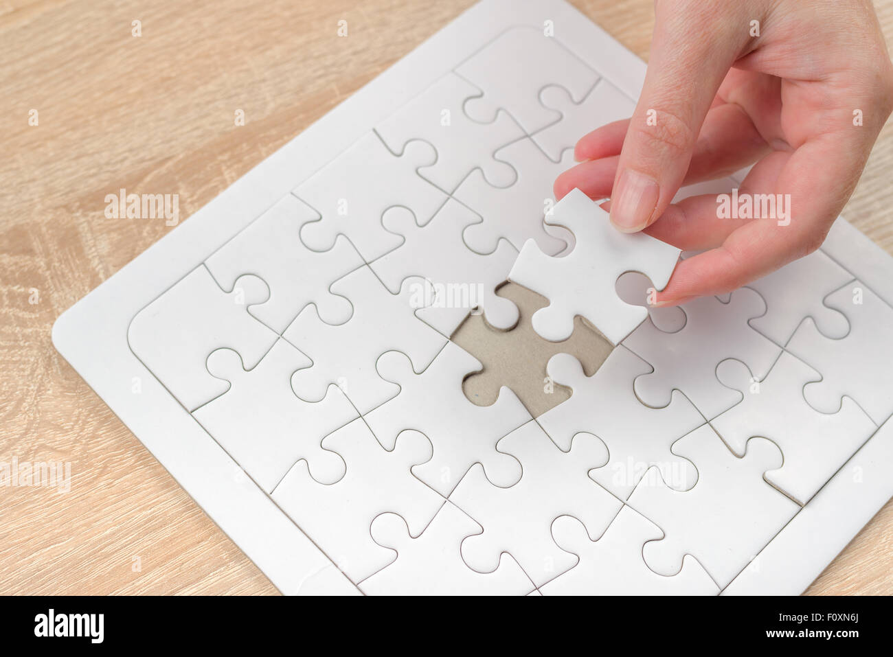 Female hand putting a missing piece and solving blank white jigsaw puzzle placed on top of old wooden oak table, close up Stock Photo