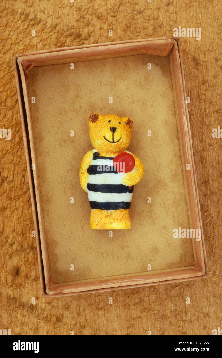 Small plaster model of smiling teddy bear with striped vest and holding red ball lying in cardboard tray Stock Photo