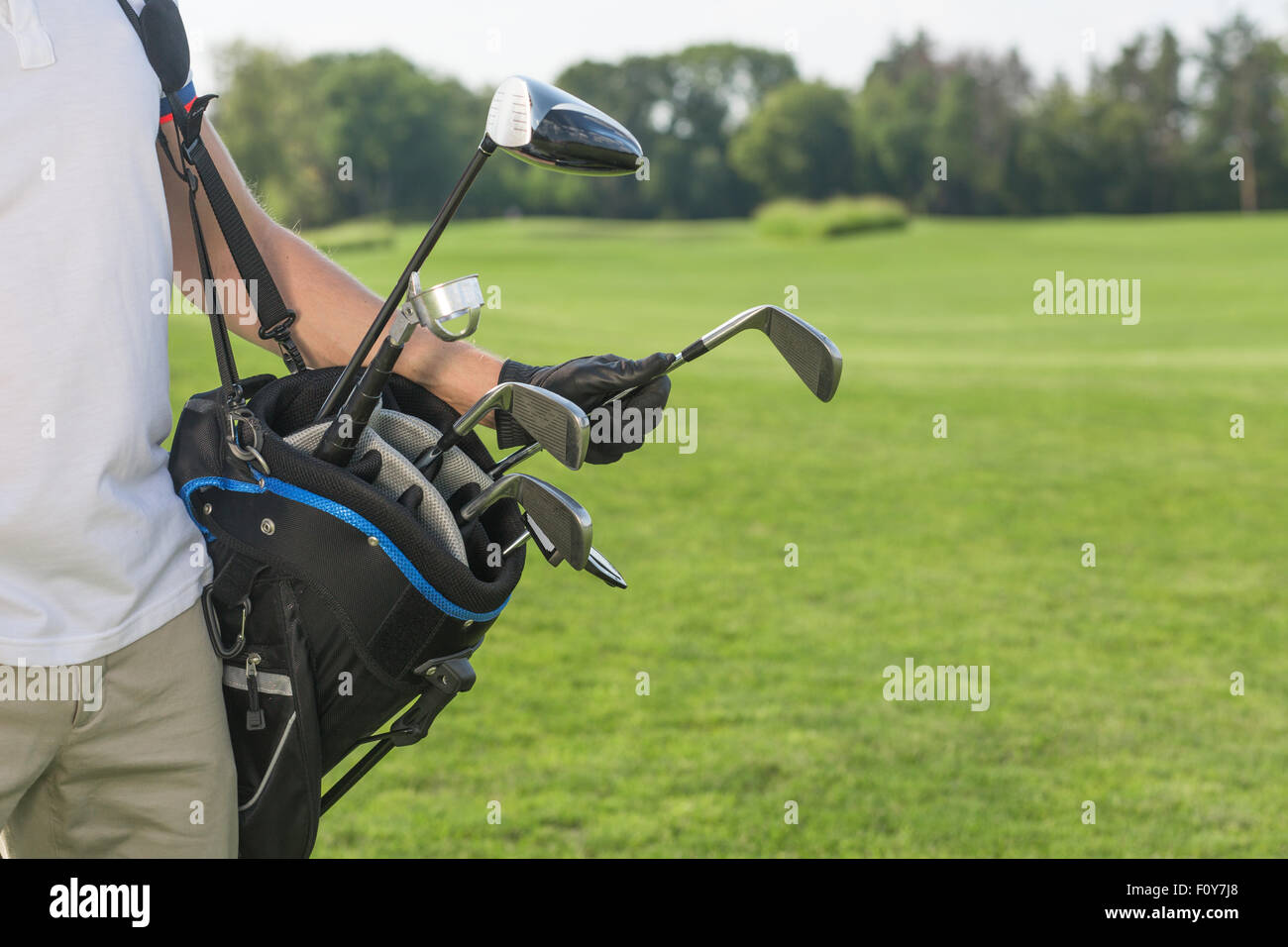 Close-up picture of golf bag hold by a golfer. Man in white T-shirt and cream trousers taking a driver to start his game over go Stock Photo