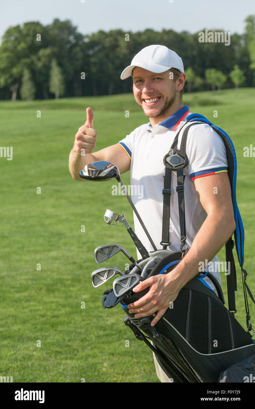 Golfer showing okay sign and smiling for the camera. Man in white trucker hat giving thumb-up and holding bag with golf drivers. Stock Photo