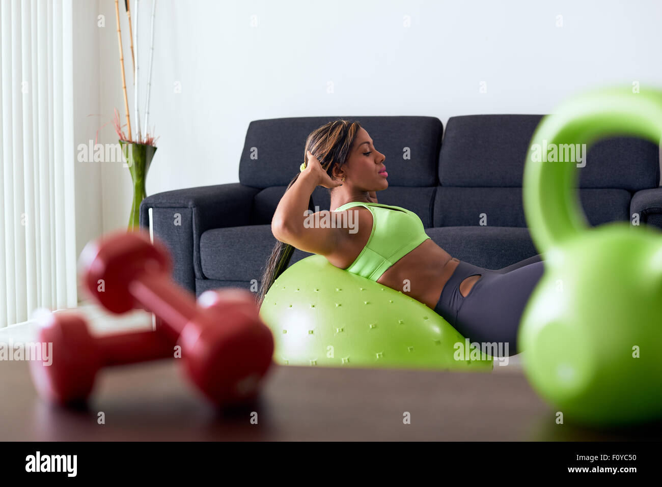 Young adult african american woman in sports clothing at home, doing domestic fitness and training abdominals on swiss ball in l Stock Photo