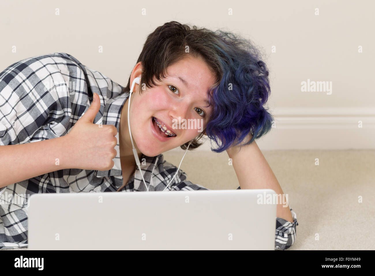 Smiling teen girl, looking forward, giving thumbs up while using computer and listening to music at home. Stock Photo