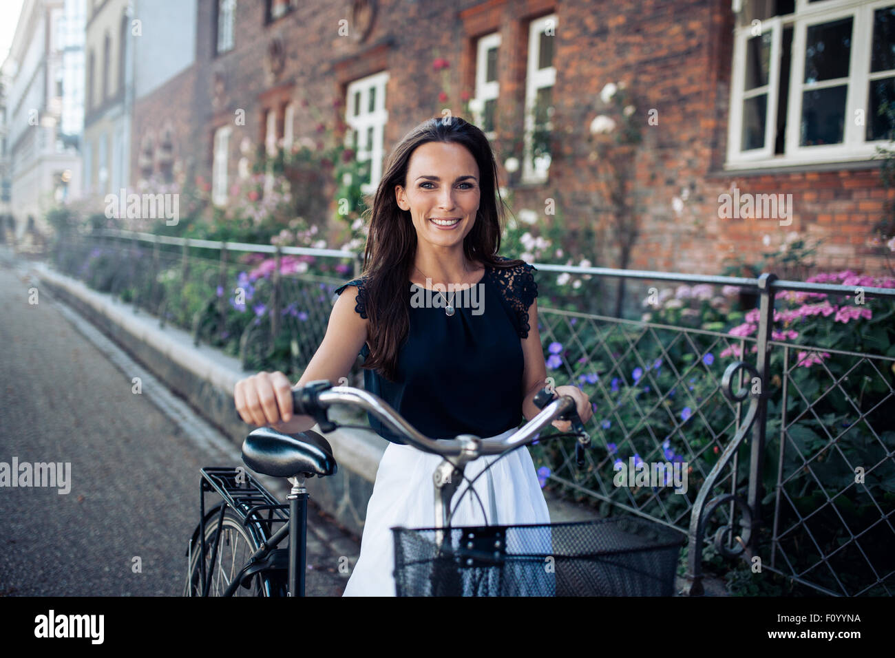 Beautiful woman with a cycle walking down the city road. Caucasian female smiling and looking at camera. Stock Photo