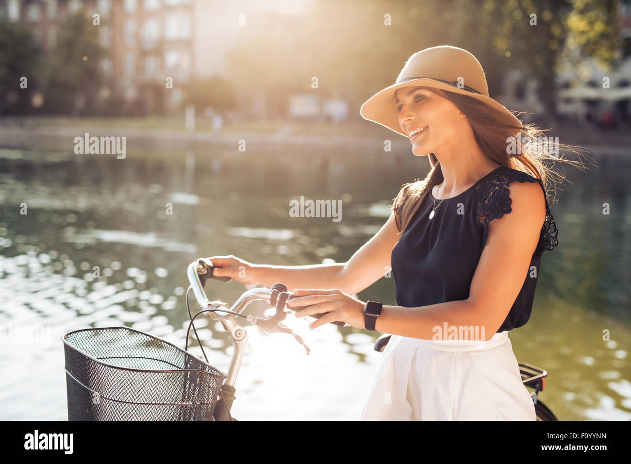 Image of beautiful woman with a bicycle at summer time near the pond at park. Female model wearing hat looking away smiling. Stock Photo