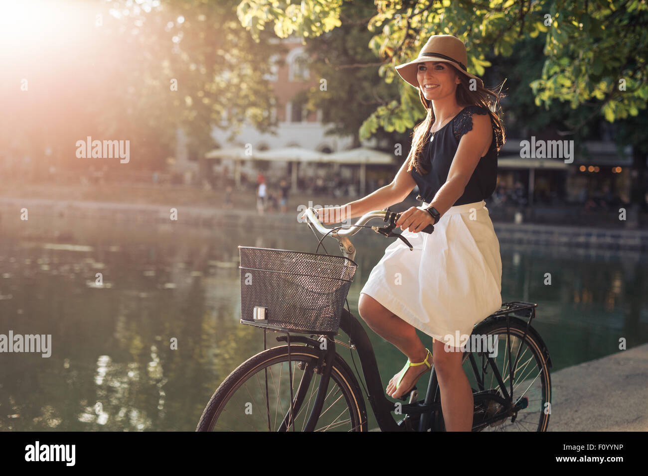 Portrait of attractive young woman riding a bicycle along a pond in city park on a summer day. Stock Photo