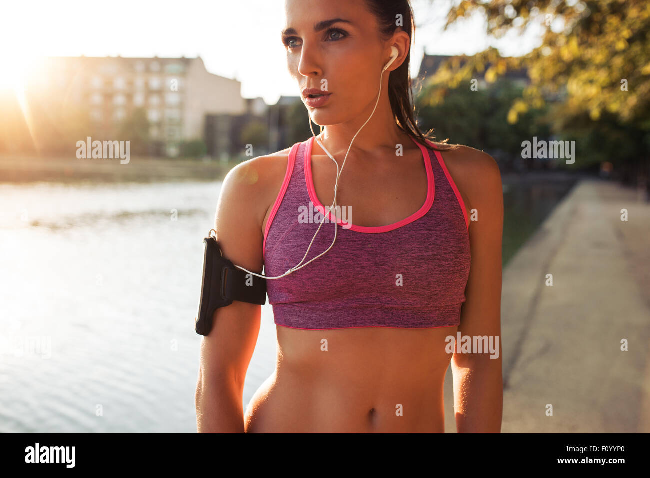 Young woman runner wearing armband and listening to music on earphones. Fit sportswoman taking a break from outdoors training. Stock Photo