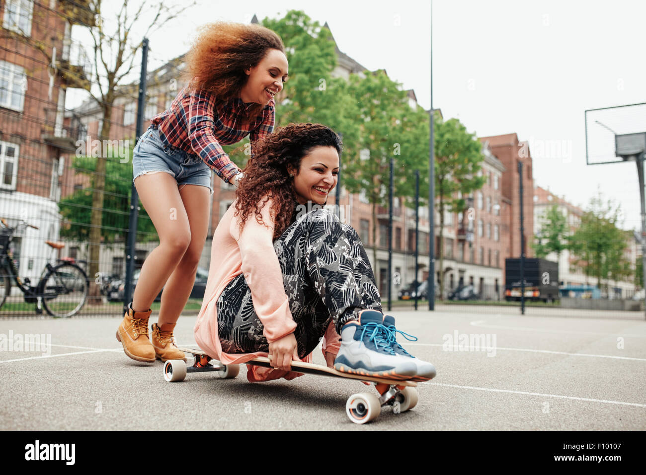 Happy young girl sitting on longboard being pushed by her friend. Young women enjoying skating outdoor. Stock Photo