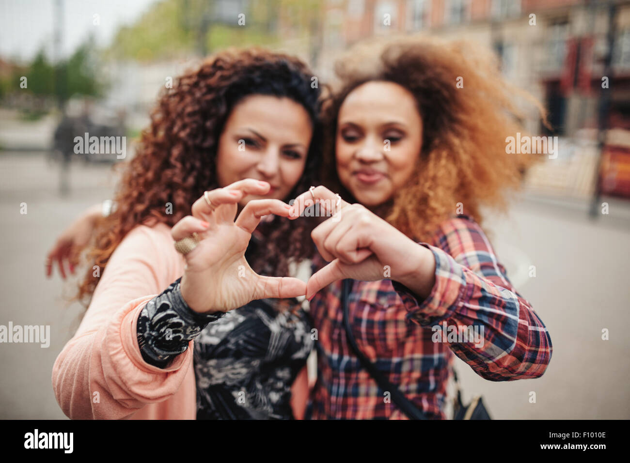 Happy young female friends making heart shape with hands and fingers. Two women standing together outdoors on city street. Focus Stock Photo