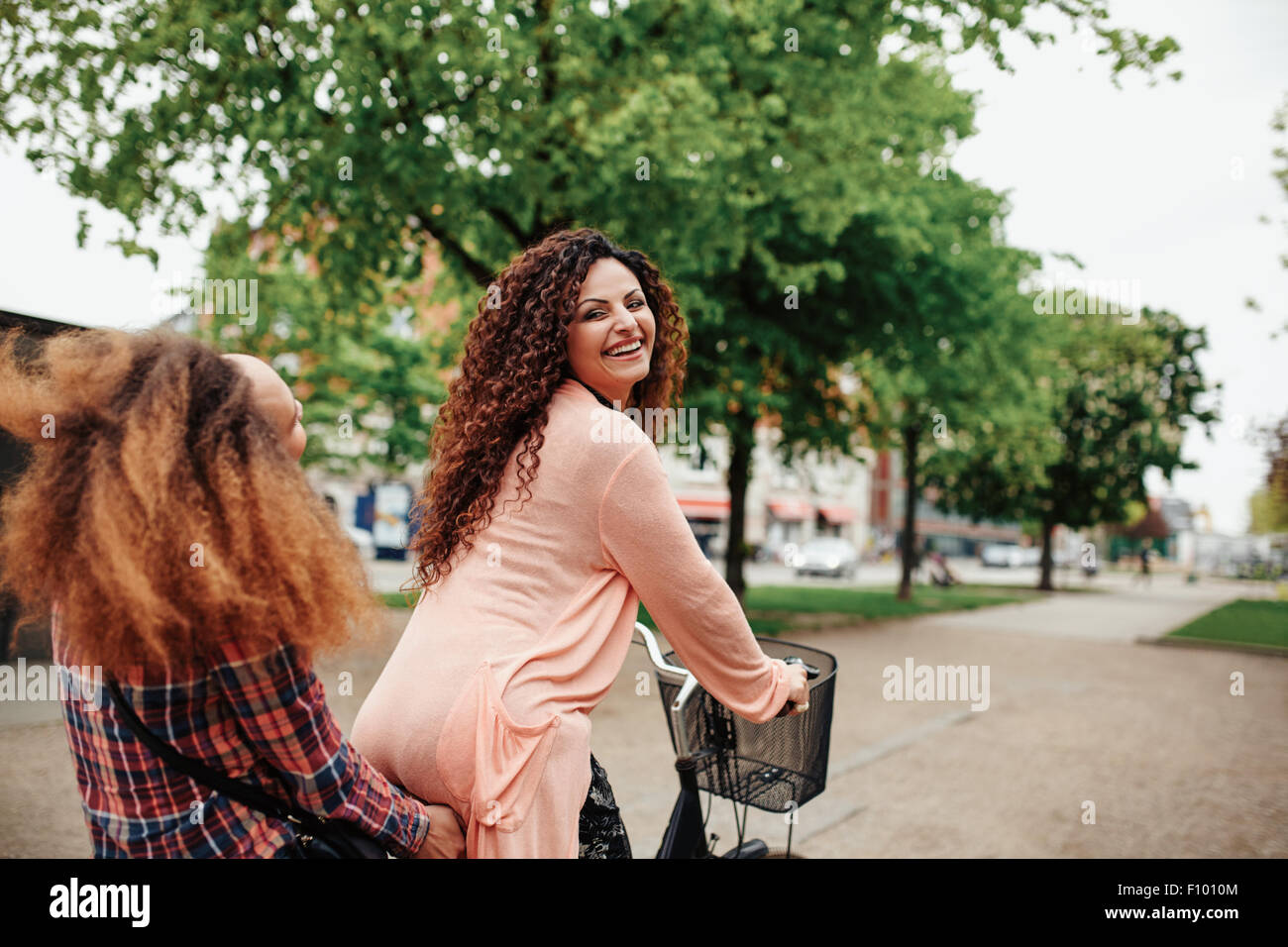 Portrait of beautiful young woman riding bicycle with her friend. Female friends having fun on cycle ride on city street. Stock Photo