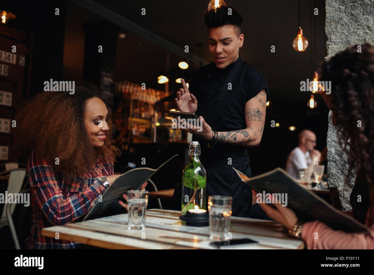 Happy young people sitting in restaurant reading menu card and giving orders to the male waiter, while waiter putting order onto Stock Photo