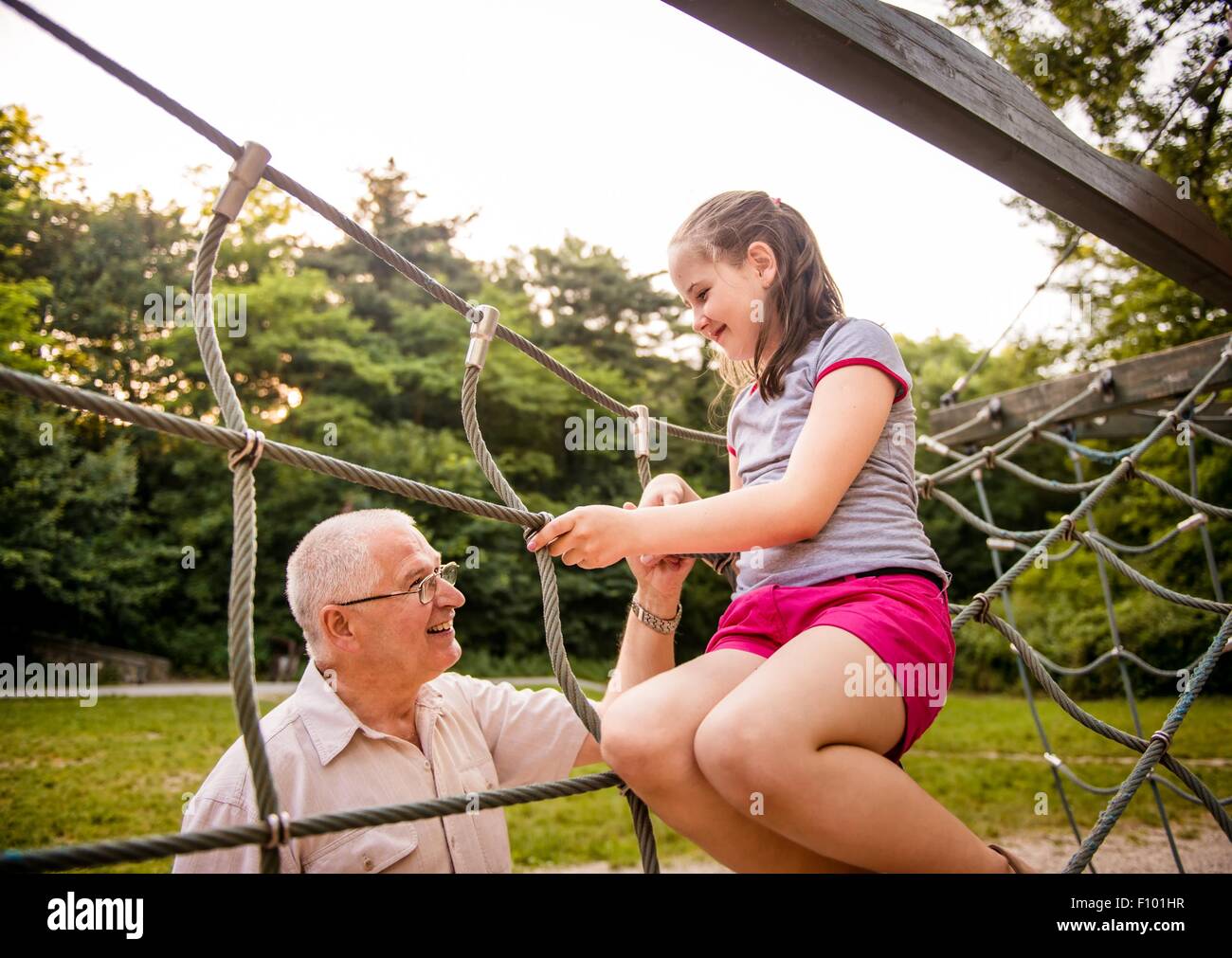 Smiling child sitting on playground net with her grandfather Stock Photo
