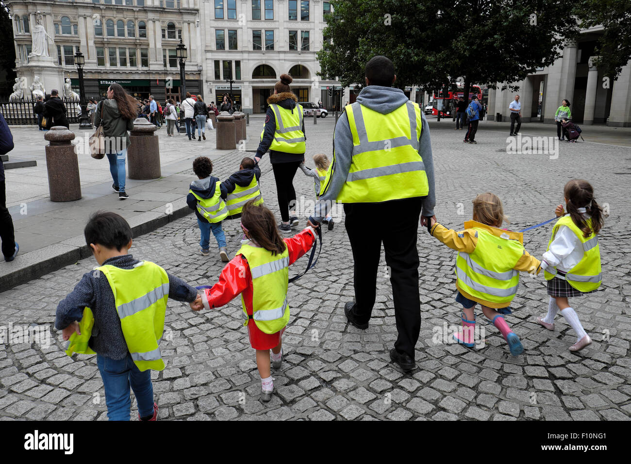 British nursery school children & childminders wearing high visibility fluorescent safety jackets hold hands on walk London street UK   KATHY DEWITT Stock Photo
