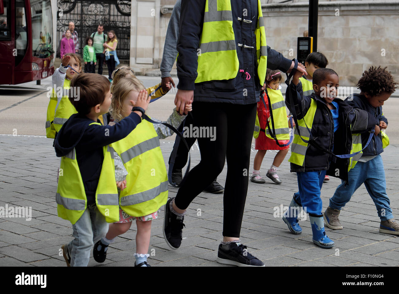 Nursery school children & childminder wearing high visibility fluorescent safety jackets hold hands on walk in a street in London UK   KATHY DEWITT Stock Photo