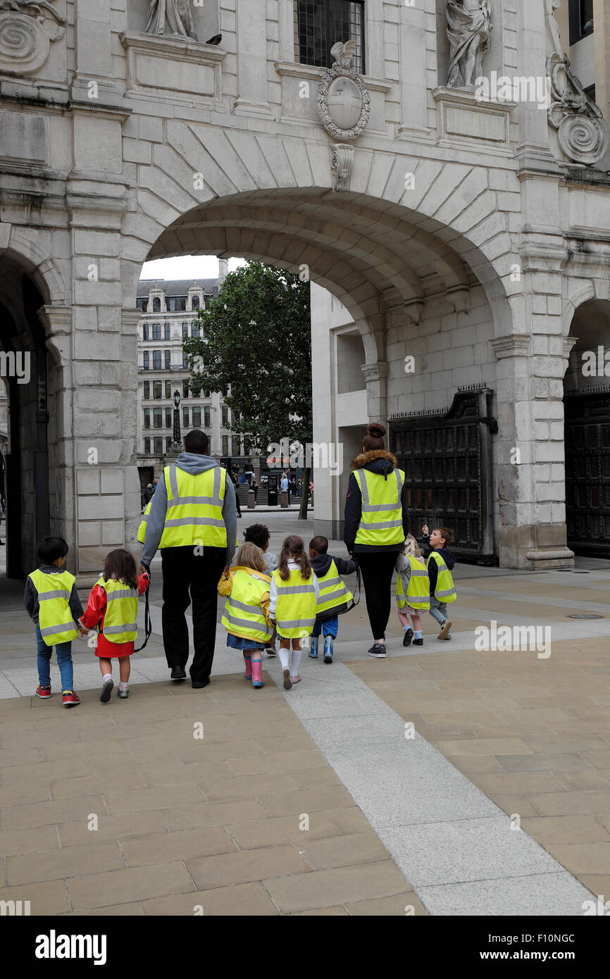 Nursery school children & child minders wearing high visibility fluorescent safety jackets hold hands on walk in a street in London UK   KATHY DEWITT Stock Photo