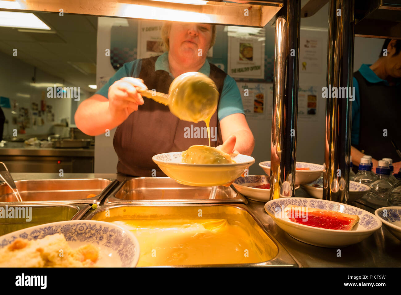 School dinners: A woman dinner lady at work pouring custard over a pudding at a school canteen cafeteria, UK Stock Photo