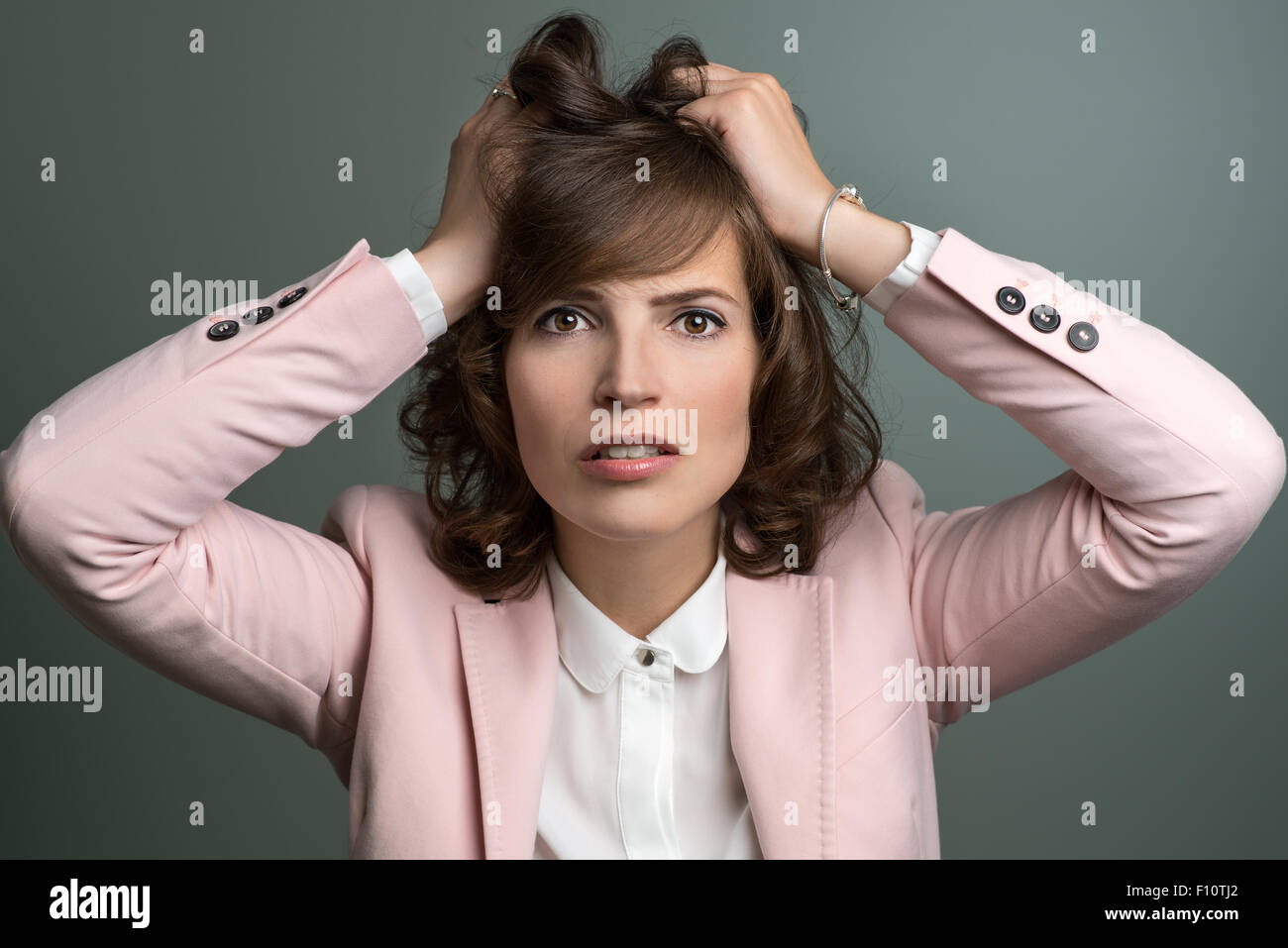 Attractive stylish young woman in a pink jacket tearing at her brown hair with her hands as she vents her frustration, over grey Stock Photo