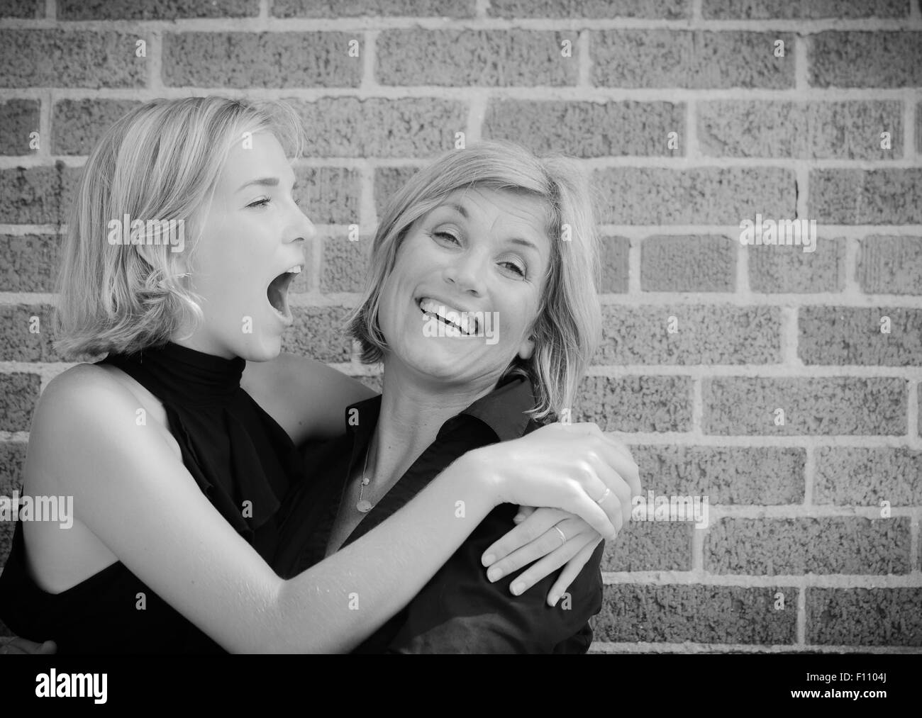 Daughter and Mother Laughing and Joking Together with Arms Around Each Other with Brick Wall in Background Stock Photo