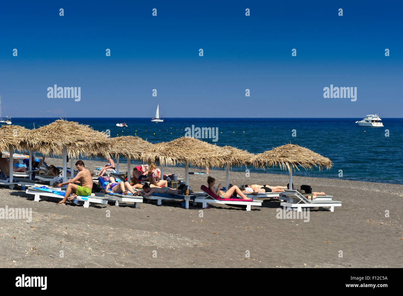 Tourists Sunbathing On The Sunny Beach Of Perissa, Santorini, Greece 