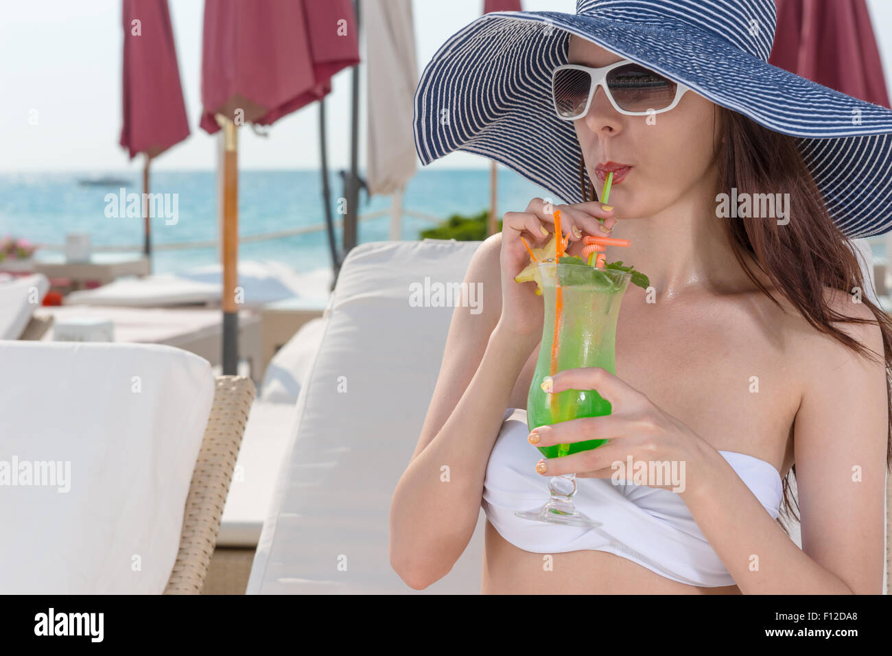 Elegant young woman in a straw sunhat and sunglasses sipping a tropical cocktail as she relaxes on a recliner chair at a seaside resort. Stock Photo