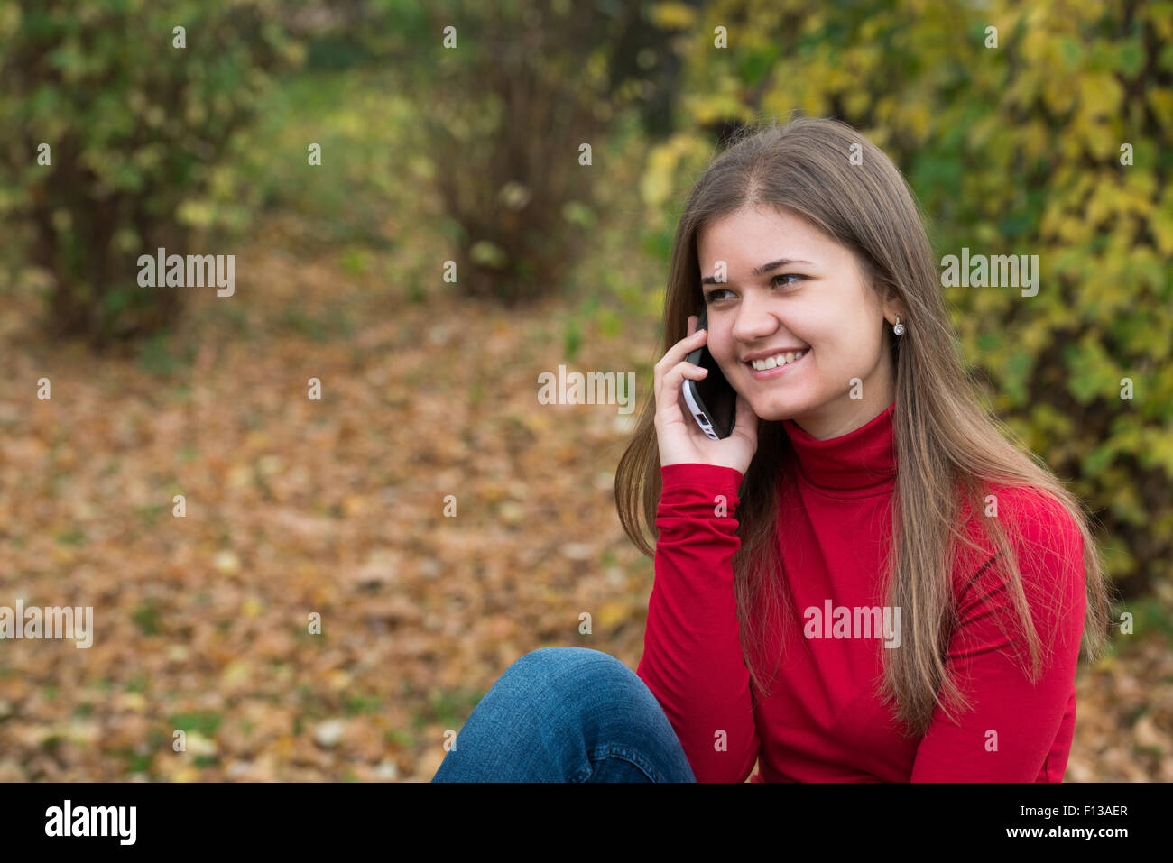 Young woman sitting  and talking on mobile phone in autumn park, in front of yellow leaves, wearing red pullover Stock Photo
