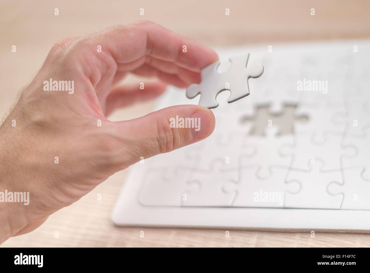 Male hand putting a missing piece and solving blank white jigsaw puzzle placed on top of old wooden oak table, close up Stock Photo