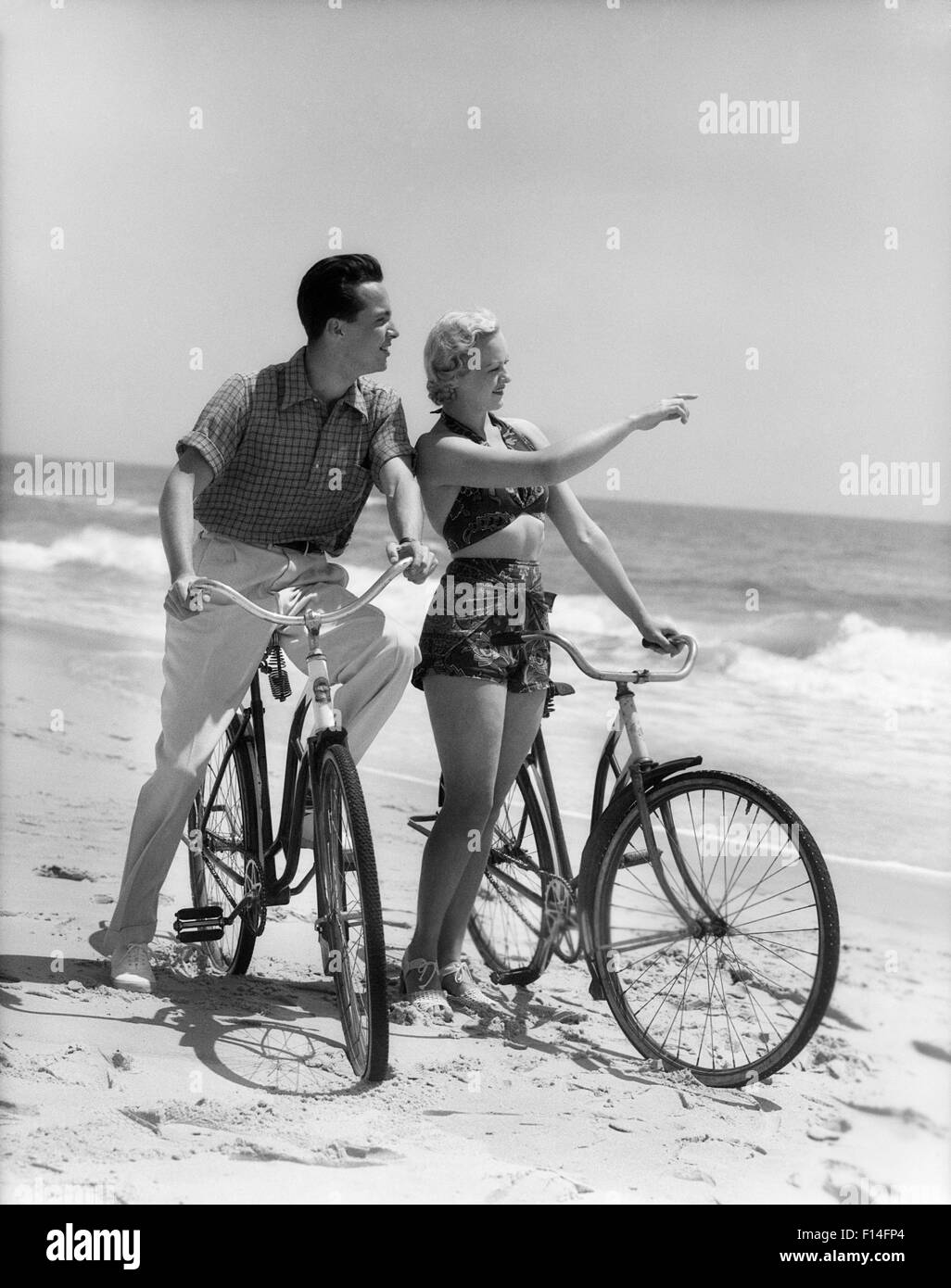 1930s Couple Man Woman Biking On Beach Woman Pointing To Horizon Stock