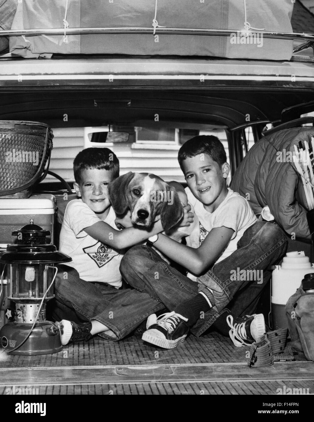 1960s TWO SMILING BOYS BROTHERS HUGGING PET DOG LOOKING AT CAMERA SITTING IN BACK OF PACKED STATION WAGON CAR READY FOR CAMPING Stock Photo