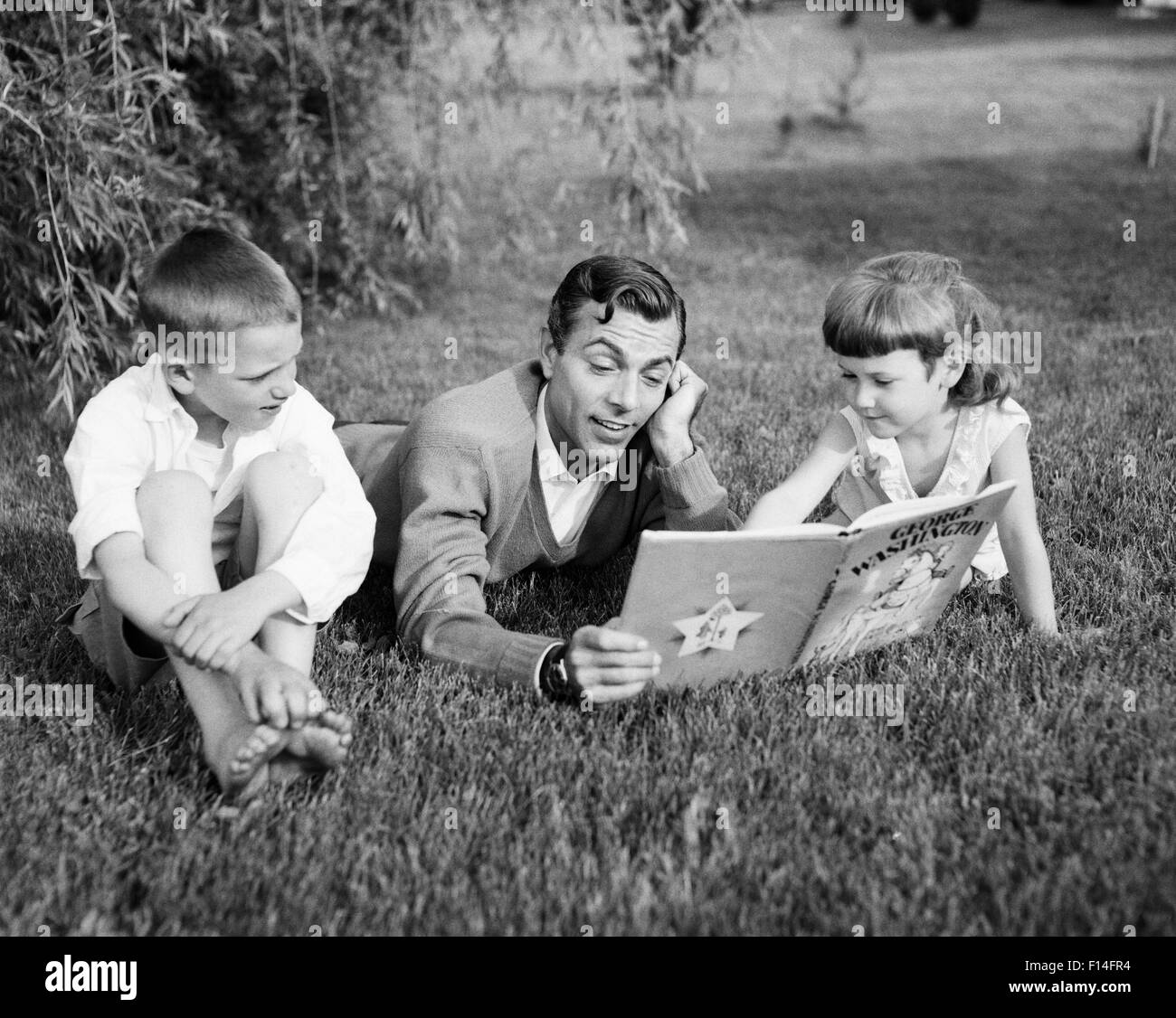 1950s MAN FATHER LYING IN THE GRASS READING BOOK TO SON AND DAUGHTER Stock Photo