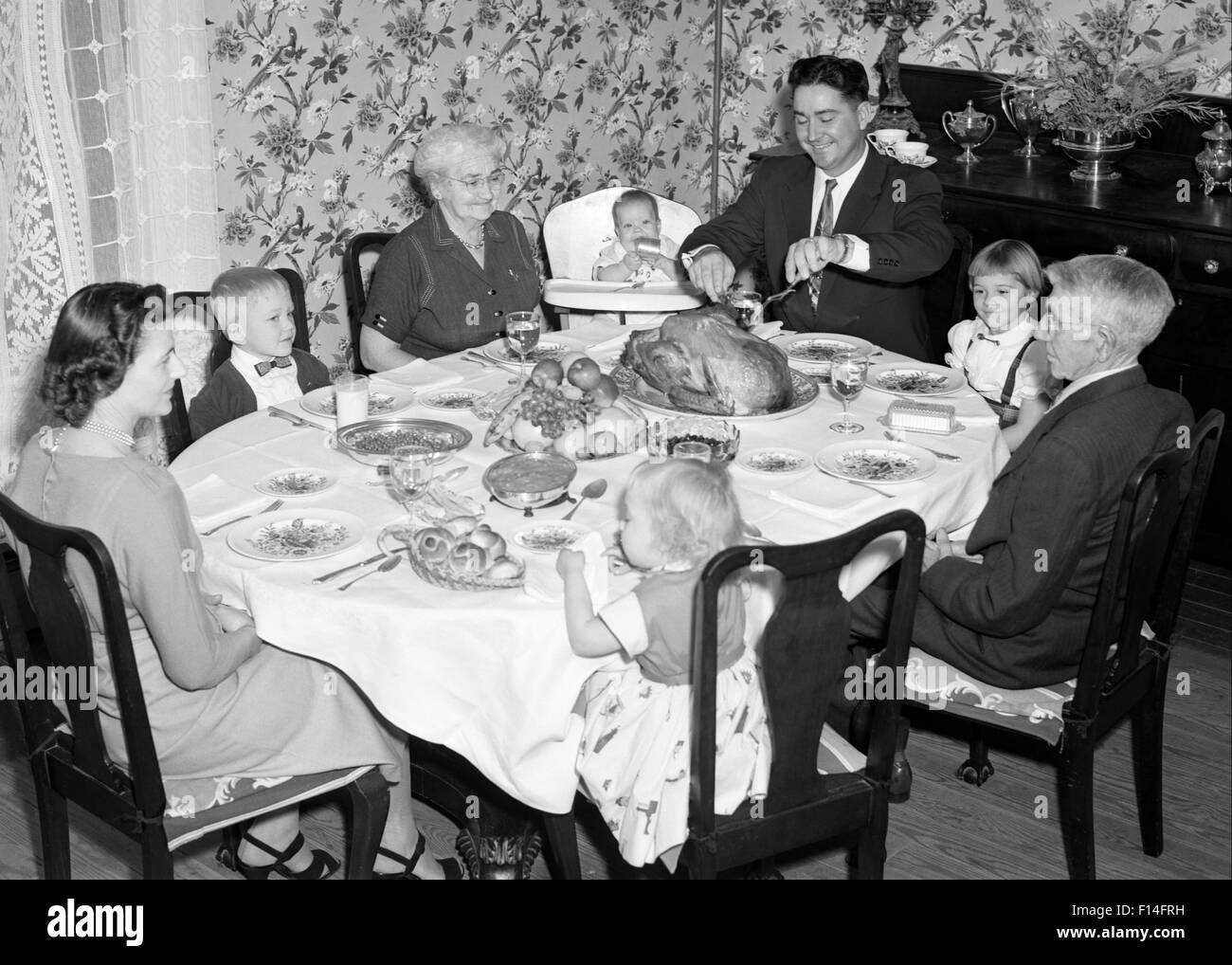 1950s THREE GENERATION FAMILY HAVING THANKSGIVING HOLIDAY MEAL IN DINING ROOM FATHER CARVING TURKEY Stock Photo