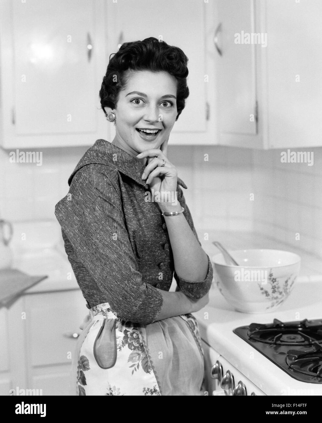 1950s SMILING BRUNETTE WOMAN HOUSEWIFE BESIDE STOVE WEARING APRON IN KITCHEN SURPRISED FACIAL EXPRESSION LOOKING AT CAMERA Stock Photo