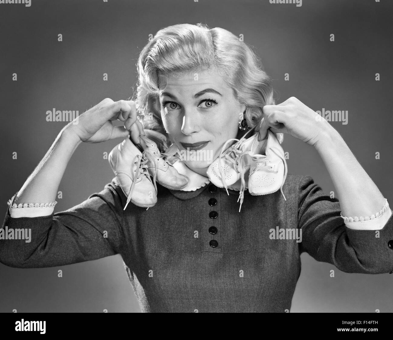 1950s PORTRAIT BLOND WOMAN HOLDING UP A PAIR OF BABY BOOTIES IN EACH HAND LOOKING AT CAMERA Stock Photo