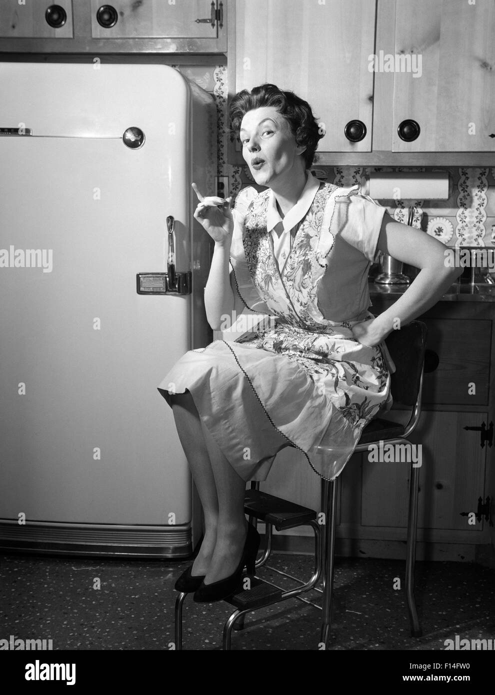 1950s HOUSEWIFE SITTING ON STOOL IN KITCHEN POINTING FINGER LOOKING AT CAMERA Stock Photo