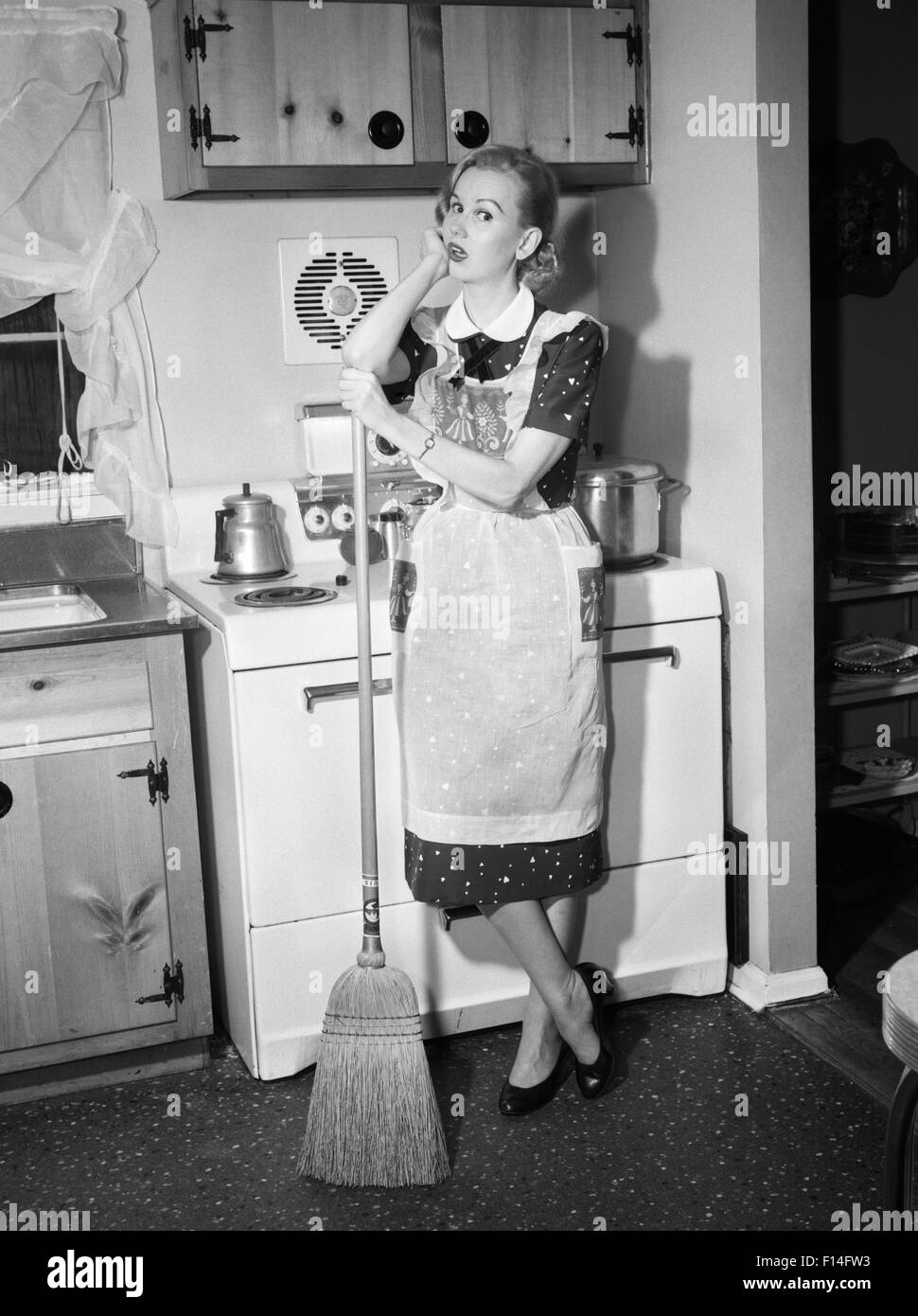 1950s TIRED HOUSEWIFE IN APRON STANDING IN FRONT OF STOVE LEANING ON BROOM LOOKING AT CAMERA Stock Photo