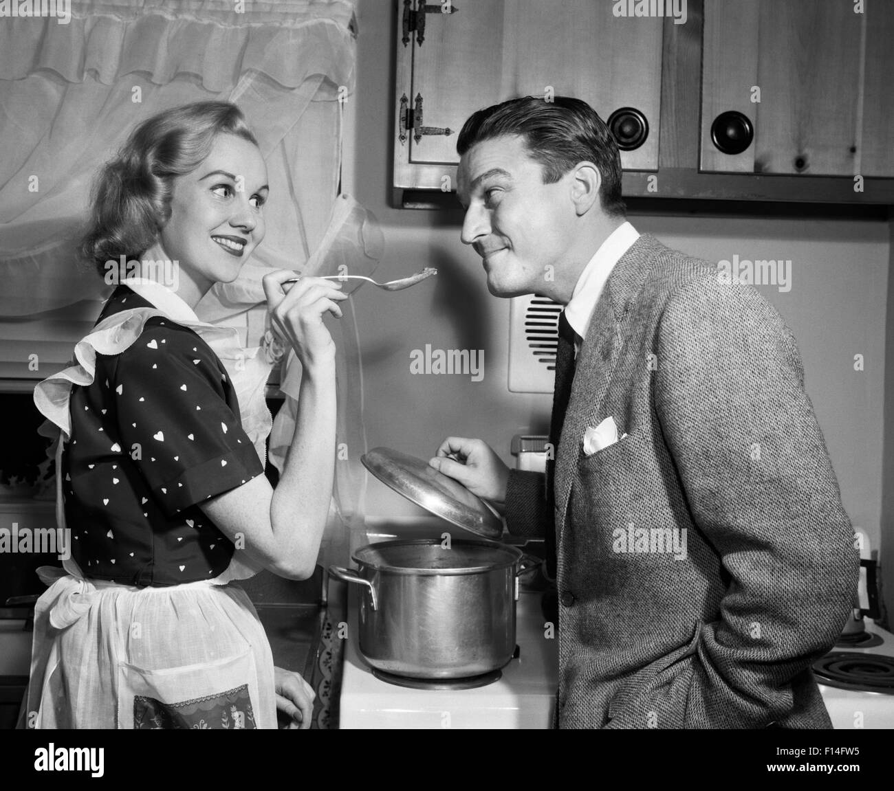 1950s SMILING HOUSEWIFE AT STOVE GIVING HAPPY HUSBAND TASTE OF HER COOKING Stock Photo