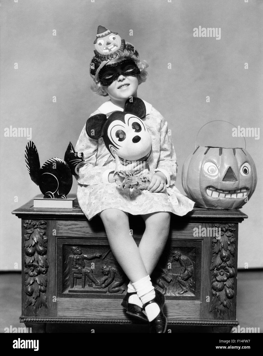 1930s LITTLE GIRL HOLDING MICKEY MOUSE TOY WEARING MASK CLOWN HAT LOOKING AT CAMERA SITTING BESIDE BLACK CAT HALLOWEEN PUMPKIN Stock Photo