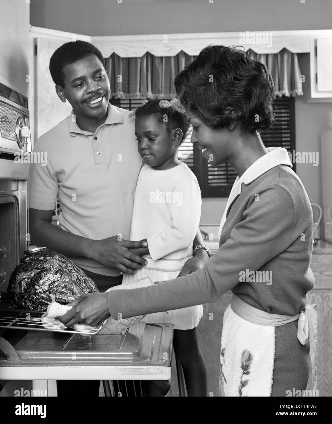1960s AFRICAN AMERICAN FAMILY IN KITCHEN FATHER AND DAUGHTER WATCHING MOTHER REMOVE ROAST TURKEY FROM OVEN Stock Photo
