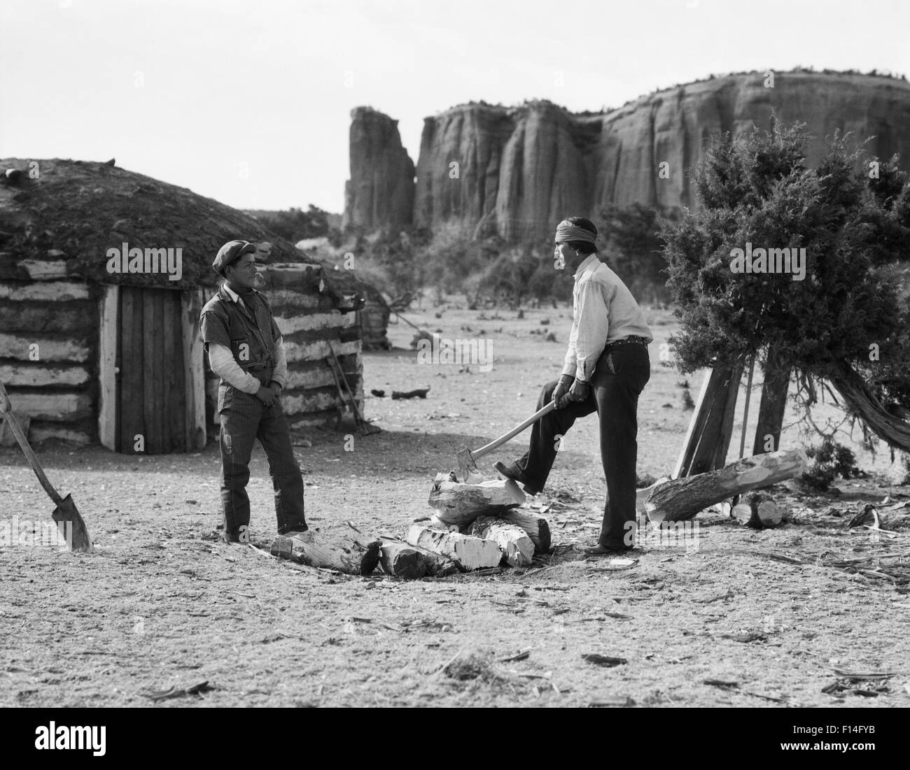 1930s TWO NATIVE AMERICAN NAVAJO MEN SPLITTING FIRE WOOD WITH AX TRADITIONAL HOGAN HOME IS IN THE BACKGROUND Stock Photo