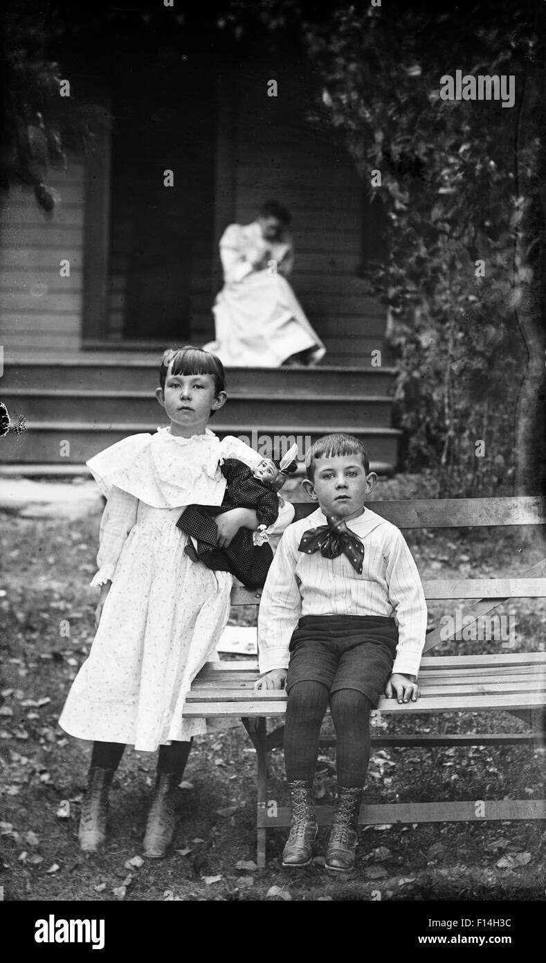 1900s PORTRAIT BOY GIRL POSING SITTING ON BENCH IN FRONT OF HOUSE LOOKING AT CAMERA WOMAN IN BACKGROUND ON PORCH Stock Photo