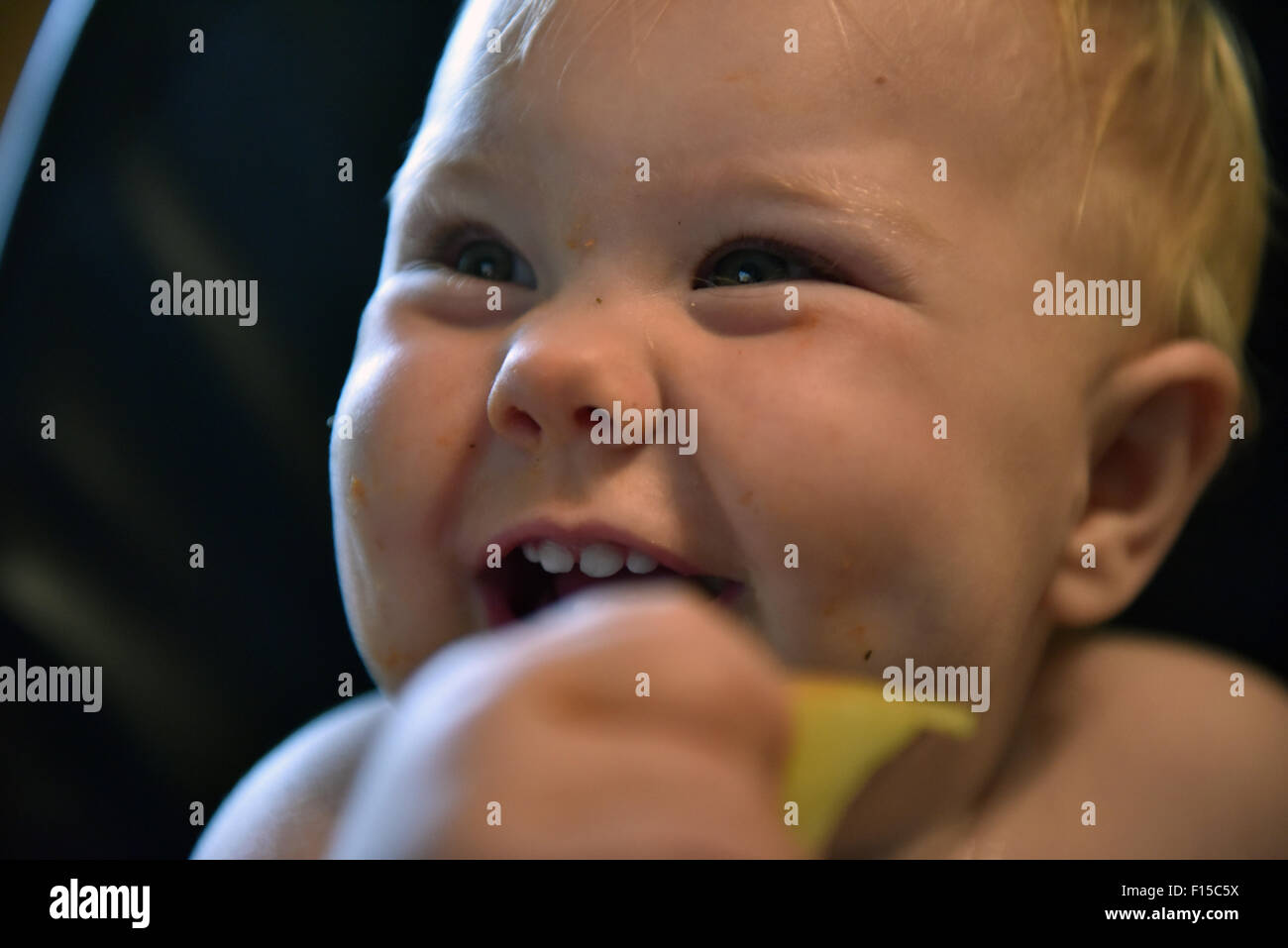 An 11 month old baby sitting in a high chair smiling with a slice of apple in her hand. Stock Photo