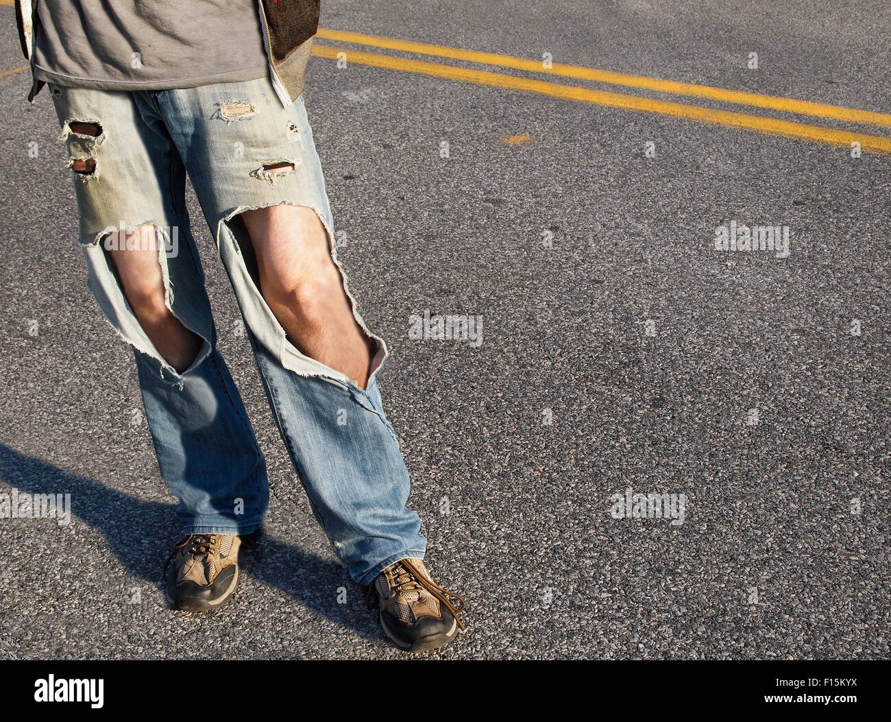 A young man, from the waist down,  in a causal t-shirt and ripped jeans with hair legs exposed stands in the road. Stock Photo