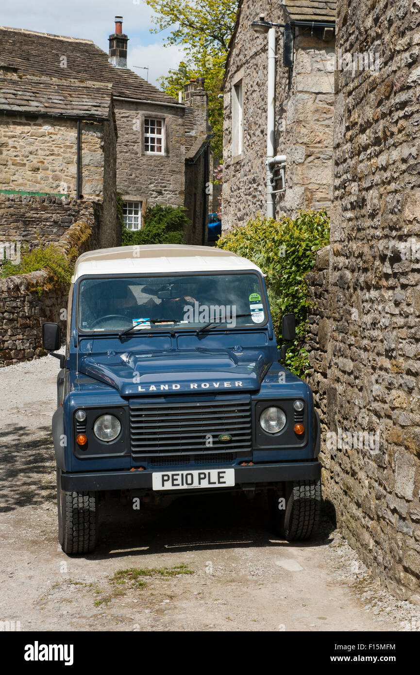Front view of clean, shiny, blue Land Rover Defender (iconic rugged 4x4 vehicle) parked on sunny Yorkshire Dales village road - Burnsall, England, UK. Stock Photo