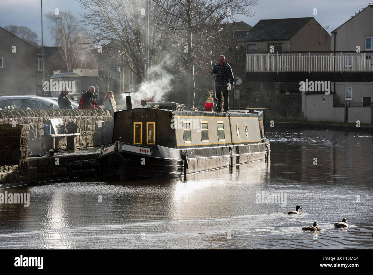Watched by people, man with brush, is standing on roof & washing moored canal boat - Leeds Liverpool Canal, Skipton, North Yorkshire England, UK. Stock Photo