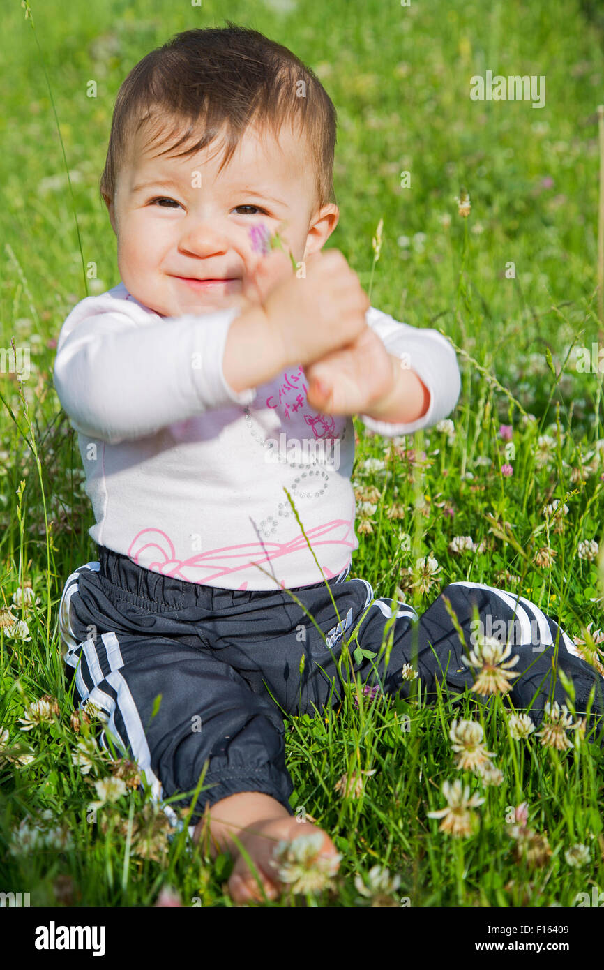 A six months old baby girl sitting on a lawn and showing a flower in her hands Stock Photo
