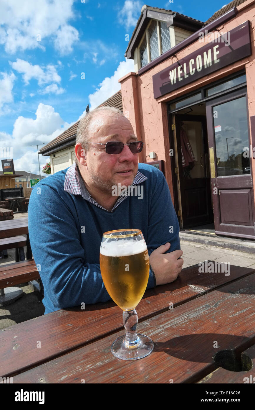 Middle-aged man sat outside a pub drinking a pint of lager with Welcome sign in background Stock Photo