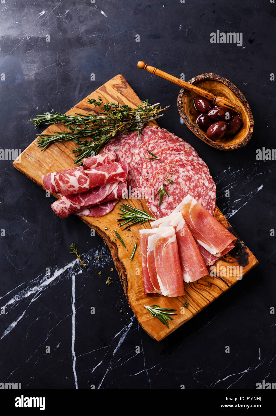 Sliced prosciutto di Parma on wooden board with salami and rosemary on black marble background Stock Photo
